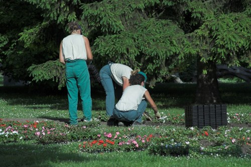 The image depicts three individuals engaged in gardening activities in a lush, well-maintained outdoor garden space. One person stands upright, wearing a sleeveless top and teal trousers, observing or preparing to work on a plant or flower bed. Another person is bent over, tending to a patch of soil or flowers, while the third is kneeling, actively planting or weeding among colorful flower beds featuring pink, red, and white blooms. The garden has a neatly trimmed grassy lawn in the foreground, and a large, dense evergreen tree provides a lush green backdrop, creating a shaded environment. The paved or gravel path area is visible near the flower beds, and a black garden rake or mat lies on the ground in the background. The overall scene suggests a professionally maintained garden, possibly in a residential or municipal setting within the UK, such as near Bracknell, allowing Garden Maintenance Archway to provide landscaping and garden care services that enhance outdoor spaces during the spring or summer months, with soft natural lighting indicating a moderate weather day.